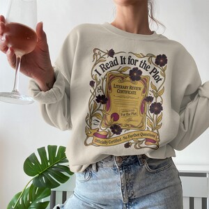 May include: Beige sweatshirt with the text "I Read It for the Plot" in a decorative design. The design includes a literary review certificate, flowers, and the phrase "Officially Certified - No Further Questions."