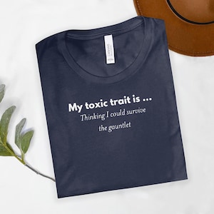 Pode incluir: Camiseta azul marinho com o texto branco: "My toxic trait is... Thinking I could survive the gauntlet". A camiseta está dobrada sobre uma superfície branca. Um chapéu marrom é visível no fundo. Feita de um material macio.