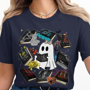 May include: Navy blue t-shirt featuring a white ghost reading a book, surrounded by various book covers with titles like "Madness" and "God of Wrath." A spider, bats, and a flower are also included in the design.