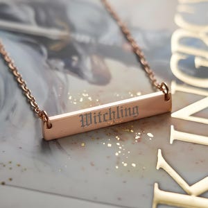 A necklace with a rectangular pendant engraved with the word "Witchling" is displayed on a surface with a book or magazine cover featuring the word "KINGDOM" in the background.
