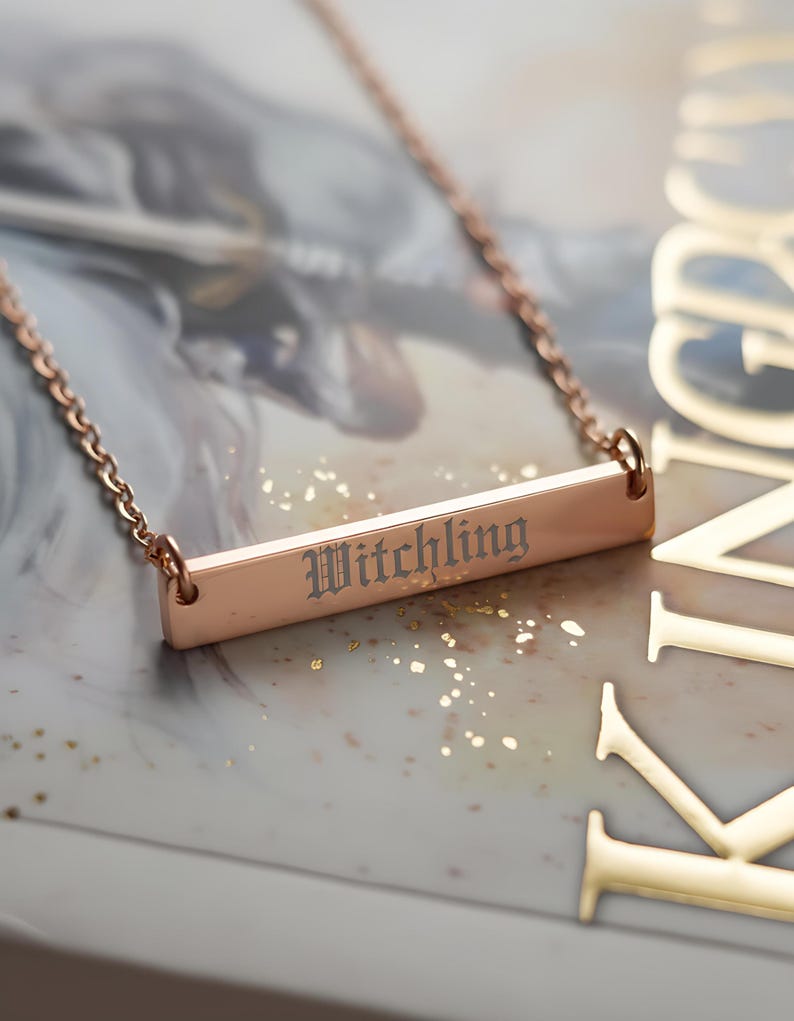 A necklace with a rectangular pendant engraved with the word "Witchling" is displayed on a surface with a book or magazine cover featuring the word "KINGDOM" in the background.