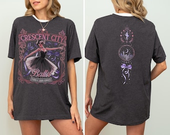 May include: Dark grey t-shirt with a white collar. The front displays a ballet-themed design with "Crescent City Ballet" and a ballerina illustration. The back has a ballet-themed design.