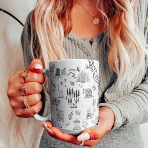 A woman with long blonde hair is holding a mug with various illustrations and text on it.