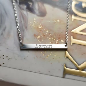 Puede incluir: Un collar plateado con un colgante rectangular grabado con el nombre "Lorcan" en cursiva. La cadena es fina y plateada. El fondo es una suave mezcla borrosa de colores, resaltando el nombre.