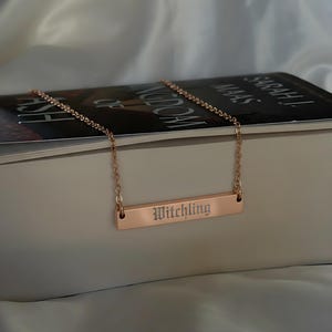 A necklace with a rectangular pendant engraved with the word "Witchling" is resting on top of a book.