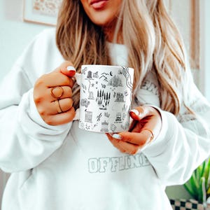 A woman with long blonde hair is holding a mug with various illustrations on it, wearing a white sweater.
