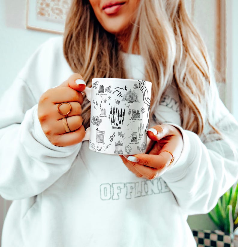 A woman with long blonde hair is holding a mug with various illustrations on it, wearing a white sweater.