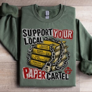 May include: Sage green sweatshirt with the text "SUPPORT YOUR LOCAL PAPER CARTEL". The graphic features a stack of yellow books chained together with a padlock. The books have titles like "Banned Thoughts" and "Knowledge is Contraband".