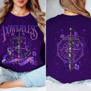 Może przedstawiać: Fioletowa bluza z nadrukiem graficznym przedstawiającym miecz, kwiaty i słowo "POWERLESS". Projekt zawiera tekst "Chcę, żeby tak było, bo ona pragnie siebie dla chcenia mnie."