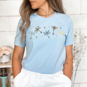 May include: Light blue t-shirt featuring a dragonfly design made of keys and stars. The dragonfly keys are in gold, blue, and yellow. The shirt is a crew neck and has short sleeves.