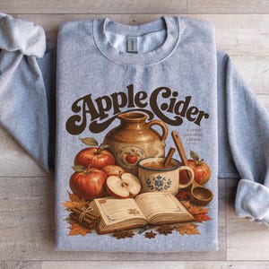 Grey sweatshirt with "Apple Cider" graphic featuring apples, a jug, and cinnamon sticks.