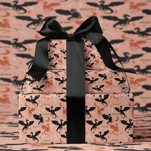 May include: A stack of wrapped gifts with a black bow. The wrapping paper has a repeating pattern of black and orange dragons, swords, and moons on a pink background. The text "DRAGON RIDER" is visible on the wrapping paper.