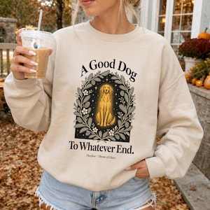 May include: A beige crewneck sweatshirt featuring a graphic of a golden dog sitting inside a botanical wreath. The text reads A Good Dog To Whatever End and Fleetfoot - Throne of Glass. The design is printed in black and gold ink.