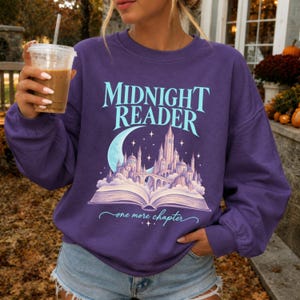 May include: A purple long-sleeve sweatshirt featuring a graphic print of an open book with a castle rising from the pages under a crescent moon and stars. The text reads Midnight Reader and one more chapter in a light blue script font.