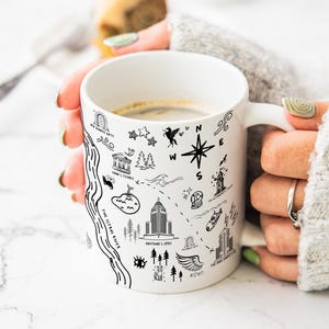 A person's hands holding a white mug with various black and white illustrations and text on it, including landmarks, symbols, and other design elements.