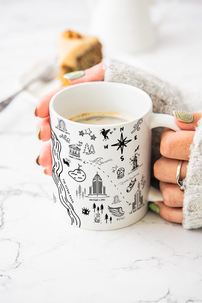 A person's hands holding a white mug with various black and white illustrations and text on it, including landmarks, symbols, and other design elements.