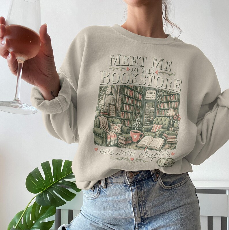 Light beige sweatshirt with "Meet Me at the Bookstore" graphic and cozy reading nook illustration.