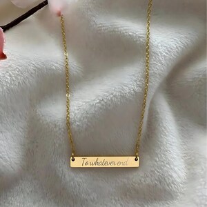 A gold-colored necklace with a rectangular pendant that says "To whatever end" engraved on it, resting on a white fabric surface.