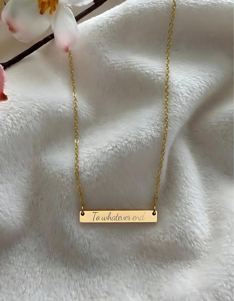 A gold-colored necklace with a rectangular pendant that says "To whatever end" engraved on it, resting on a white fabric surface.