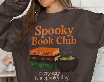 Puede incluir: Una sudadera marrón oscuro con la frase "Spooky Book Club" en naranja. Debajo, tres libros apilados con los títulos "Witchcraft", "Ghost Stories" y "Monsters". Una caldera con líquido verde y globos oculares flotantes junto a los libros. El texto "every day is a spooky day" está debajo.