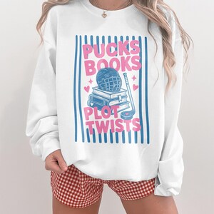 May include: A white crewneck sweatshirt featuring a graphic print with blue vertical stripes. The design includes the pink text "PUCKS BOOKS" and "PLOT TWISTS" surrounding a stack of blue books, a hockey helmet, a hockey stick, and small hearts.