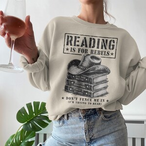 Könnte beinhalten: Beiges Sweatshirt mit dem Text "READING IS FOR REBELS" in einem schwarzen Rechteck. Darunter eine Grafik eines Cowboyhuts auf einem Stapel Bücher. Der Text "DON'T FENCE ME IN (I'M TRYING TO READ)" befindet sich darunter.