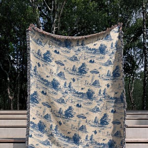 May include: A beige and blue throw blanket with a toile pattern featuring UFOs, trees, houses, and animals. The blanket has a fringed edge and is displayed outdoors.