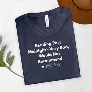 Może przedstawiać: Granatowa koszulka z białym napisem "Reading Past Midnight - Very Bad, Would Not Recommend". Koszulka ma również cztery białe gwiazdki. Brązowy kapelusz jest widoczny w tle.