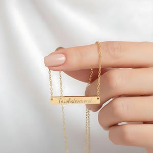 A person's hand holding a gold necklace with a rectangular pendant that says "To whatever end".