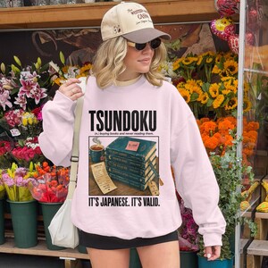 Pink sweatshirt with "Tsundoku" text and stack of unread books.