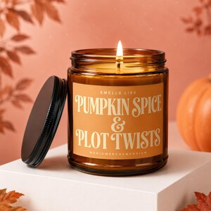 May include: A lit candle in an amber glass jar with a black lid. The label is orange with white text that reads: SMELLS LIKE PUMPKIN SPICE & PLOT TWISTS MAGICMERCHEMPORIUM. The candle is positioned on a white surface with autumn leaves nearby.