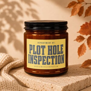 May include: A brown glass candle jar with a black lid and a yellow rectangular label. The label features the text DEPARTMENT OF: PLOT HOLE INSPECTION and MAGICMERCHEMPORIUM in dark blue block lettering centered on the front of the container.