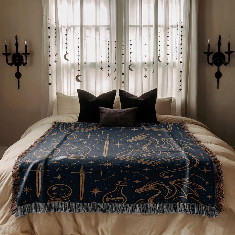 a bedroom with a large bed covered in a blue and gold blanket featuring a dragon and star motif. The bed is flanked by two black wall sconces, and the window behind the bed is decorated with white curtains and star-shaped string lights.