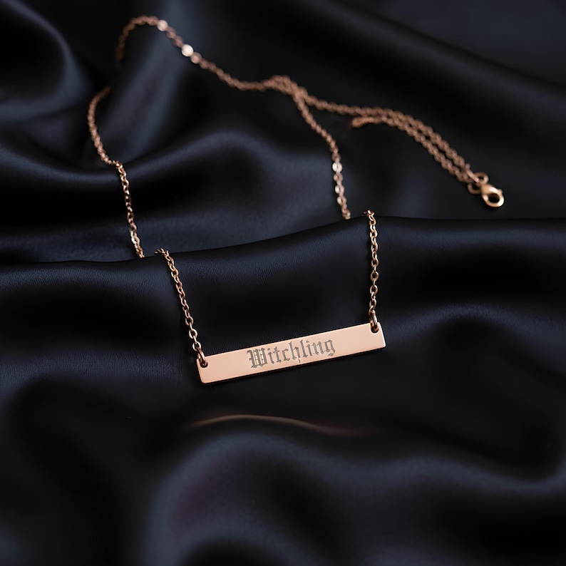 A gold-colored necklace with a rectangular pendant engraved with the word "Witchling" is displayed on a dark fabric background.