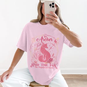 Könnte beinhalten: Hellrosa T-Shirt mit einer rosa Fuchsgrafik in einem Kreis. Der Text "the Archer" und "AND THE FOX The Magnificent North" sind rosa gedruckt. Das T-Shirt hat eine lockere, übergroße Passform.