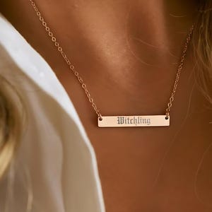 A close-up of a person wearing a gold necklace with a rectangular pendant engraved with the word "Witch".