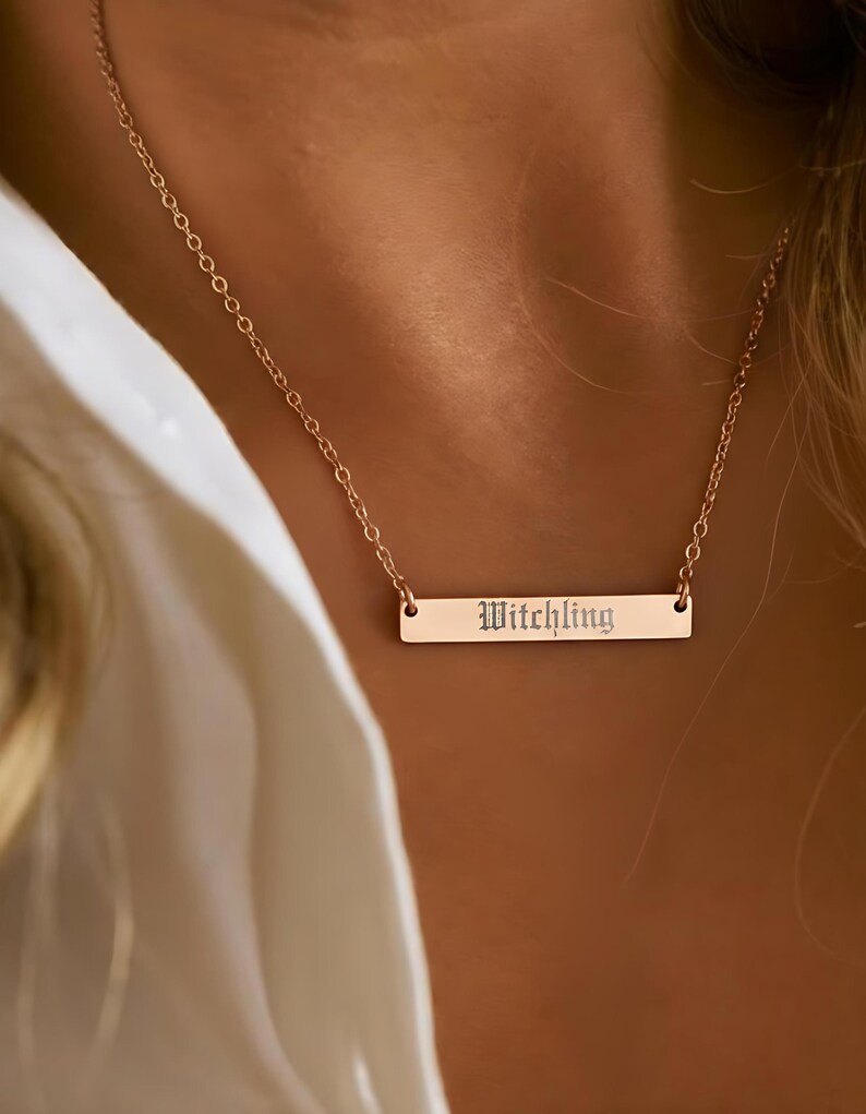 A close-up of a person wearing a gold necklace with a rectangular pendant engraved with the word "Witch".