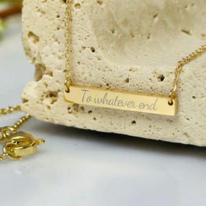 A gold-colored necklace with a rectangular pendant that says "To whatever end" engraved on it, resting on a white surface.