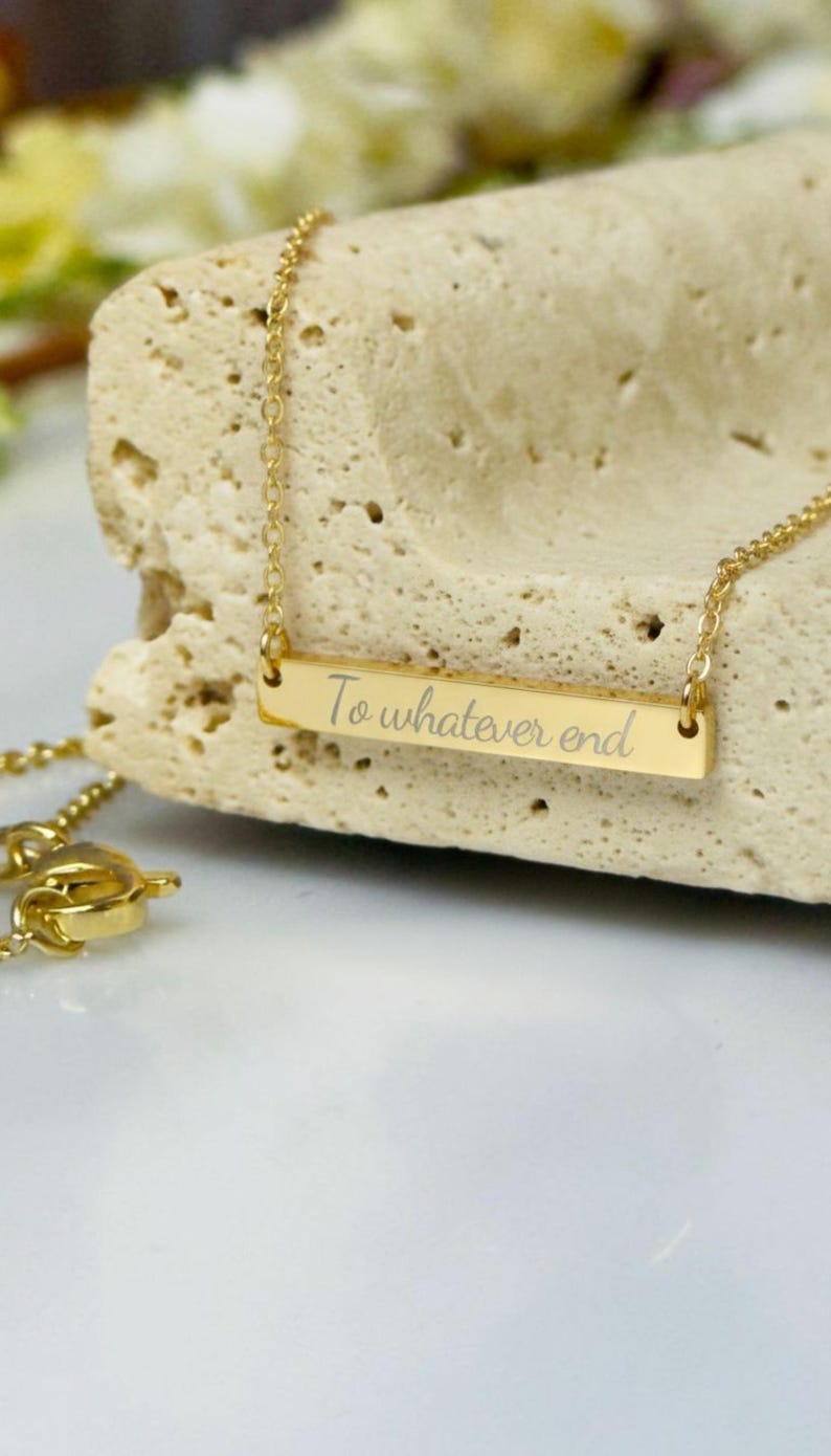 A gold-colored necklace with a rectangular pendant that says "To whatever end" engraved on it, resting on a white surface.