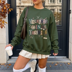 Op de afbeelding: Een olijfgroene sweatshirt met de tekst "VALKYRIE IN TRAINING" in een kleurrijk, speels lettertype. Het ontwerp bevat afbeeldingen van een koffiekopje, boeken en sterren. De sweatshirt maakt deel uit van een casual kledingcollectie.