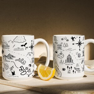 Two white mugs with black illustrations and text on them, placed on a white surface with a slice of lemon in front of them.