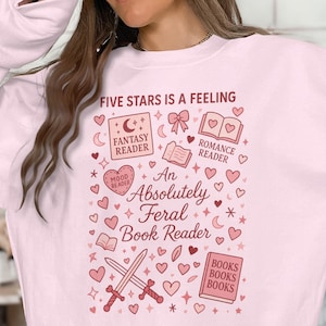 May include: Light pink sweatshirt with a graphic design featuring the text "FIVE STARS IS A FEELING" and "An Absolutely Feral Book Reader." The design includes hearts, books, swords, and the words "Fantasy Reader" and "Romance Reader."