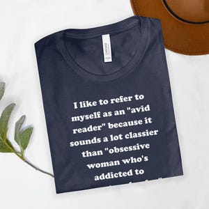 May include: Navy blue t-shirt with white text: "I like to refer to myself as an 'avid reader' because it sounds a lot classier than 'obsessive woman who's addicted to". The shirt is made of soft material.