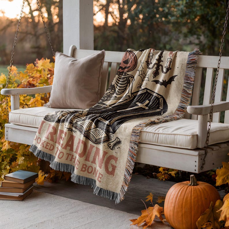 a porch with a white wooden bench, a pumpkin, and a blanket with a skeleton design. The blanket is draped over the bench, and there are books and a pumpkin nearby.