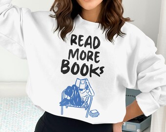 May include: White sweatshirt with the text "READ MORE BOOKS" in black. A blue illustration shows a person reading a book while seated in a chair. The sweatshirt is a casual style.