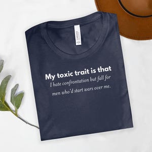 May include: Navy blue t-shirt with the text "My toxic trait is that I hate confrontation but fall for men who'd start wars over me." The shirt is made of a soft material and has a crew neck.