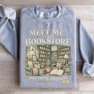 Gray sweatshirt with "Meet Me at the Bookstore" and books in a cozy setting.