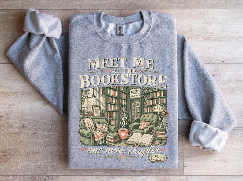 Gray sweatshirt with "Meet Me at the Bookstore" and books in a cozy setting.