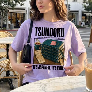 Purple t-shirt with "Tsundoku" text and stack of books graphic.