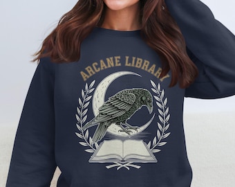 May include: Navy blue sweatshirt with a graphic featuring a raven perched on a crescent moon above an open book. The text "ARCANE LIBRARY" arches above the raven. The design is in shades of gray and tan.